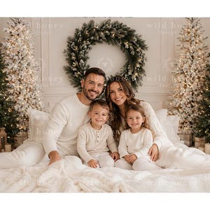 Puede incluir: Una familia de cuatro personas con camisas de manga larga y pantalones a juego de color crema, sentados juntos en una cama blanca. El fondo muestra dos árboles de Navidad iluminados y una gran corona de pino con el texto wild honey designs.