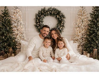 May include: A family of four wearing matching cream-colored long-sleeved shirts and pants, sitting together on a white bed. The background features two lit Christmas trees and a large evergreen wreath on the wall, with the text wild honey designs.
