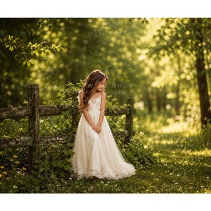 May include: A floor-length, off-white lace dress with thin spaghetti straps and a fitted bodice, worn by a child standing in a sunlit outdoor setting near a wooden fence. The dress features intricate floral patterns and a flowing, full skirt.