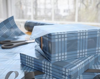 May include: Two gift boxes wrapped in blue and gray plaid wrapping paper. A roll of matching paper and scissors are visible in the background. A gray ribbon is draped across the wrapping surface. The boxes are stacked on top of each other.