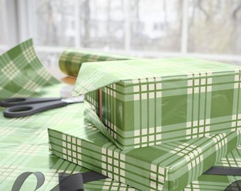 May include: Two gift boxes wrapped in green plaid wrapping paper with a white grid pattern. A roll of matching paper, scissors, and a dark gray ribbon are also visible, suggesting a theme of gift wrapping.