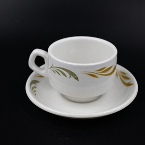Puede incluir: Juego de taza y platillo de té de cerámica blanca. La taza y el platillo presentan un diseño decorativo de hojas en oro y verde oliva. La taza tiene un asa y se apoya en un platillo a juego. El juego está sobre un fondo negro.