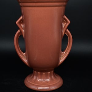 Op de afbeelding: Een terracottakleurige keramische vaas met twee handvatten. De vaas heeft een klassieke, urn-achtige vorm met een geflute voet.