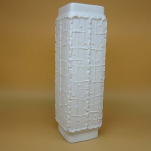 May include: A tall, white ceramic vase with a textured, grid-like pattern. The rectangular vase has a square top and base, and the surface features a raised, dripped design. The vase is a decorative home decor item.
