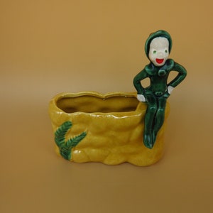 May include: A green ceramic figurine of a smiling elf sitting on a yellow ceramic planter with green leaves. The planter is shaped like a rock.