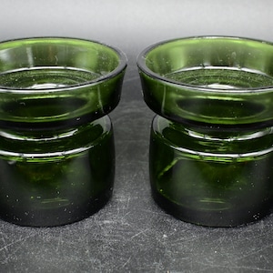 Op de afbeelding: Twee groene glazen kaarsenhouders met een brede, platte bovenkant en een cilindrische basis. De houders zijn gemaakt van dik glas en hebben een licht getextureerd oppervlak.