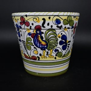 May include: A white ceramic planter with a green, yellow, blue, and red rooster design. The planter has a wide rim and a rounded base.