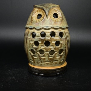 May include: A ceramic owl lamp with a brown and green glaze. The owl has a round body with cut-out holes and a small base.