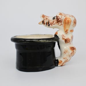 Peut inclure: Un pot en céramique en forme de chapeau haut de forme noir avec une figurine de chien tenant le côté. Le chien est blanc avec des taches brunes et a une oreille brune.