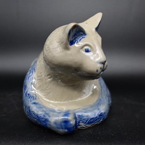 Puede incluir: Una figurita de cerámica de un gato con cuerpo blanco y detalles azules y blancos. El gato está sentado con la cabeza girada hacia un lado.