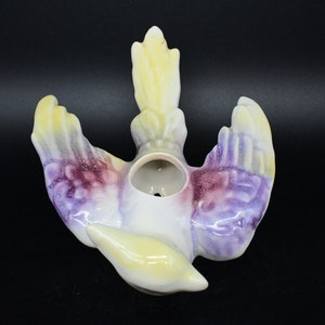 Puede incluir: Un macetero de pared de cerámica con forma de pájaro con detalles pintados en amarillo, morado y blanco. El pájaro tiene las alas extendidas y la cabeza echada hacia atrás. Hay un agujero en la parte superior de la cabeza del pájaro para plantar.