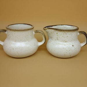 Może przedstawiać: Zestaw ceramiczny do śmietanki i cukru w kolorze białym z brązową, kropkowaną glazurą i brązowymi obwódkami.