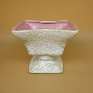 May include: A white ceramic planter with a pink glazed interior. The planter has a square top and a pedestal base with a textured surface.
