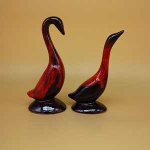 Puede incluir: Dos figuritas de ganso de cerámica con un esmalte rojo y negro. Los gansos están de pie sobre una superficie marrón claro.