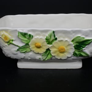 以下が含まれることがあります： 長方形の白い陶器製のプランターで、浮き彫りの花柄が施されています。プランターには、黄色い中心を持つ白い花が3つと緑の葉が描かれています。