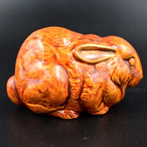 Puede incluir: Figura de conejo de cerámica en tonos naranja y marrón. El conejo está en posición sentada con las orejas hacia atrás. La figura tiene un acabado brillante y una textura detallada, lo que sugiere un artículo hecho a mano.
