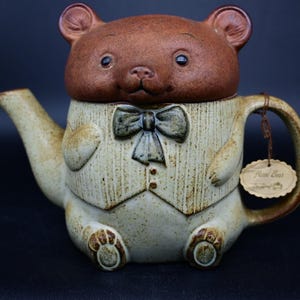 Puede incluir: Una tetera de cerámica con forma de oso que lleva una pajarita. La tetera es de color marrón y beige con un esmalte moteado. La cara del oso es marrón y tiene una expresión linda. La tetera tiene un pico, una asa y una tapa. La tetera tiene una etiqueta que dice "Home Sweet Home".