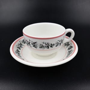 May include: White teacup and saucer set with a pink rim and a black and white floral design. The teacup has a handle and sits on a matching saucer. The set is on a black background.