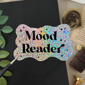 May include: A holographic sticker with the text "Mood Reader" is held by tweezers. The sticker has a glittery, iridescent finish and a scalloped edge. A black notebook, stationery, and decorative items are in the background.