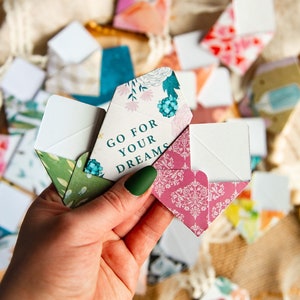 K&ouml;nnte beinhalten: Eine Hand h&auml;lt einen Satz bunter Papierumschl&auml;ge mit einem floralen Design. Der oberste Umschlag tr&auml;gt den Text "Go for your dreams".