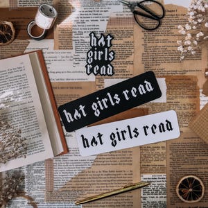 May include: Two black and white bookmarks with the text "hot girls read" in a gothic font. The bookmarks are on a table with a book, dried flowers, and other decorative items.