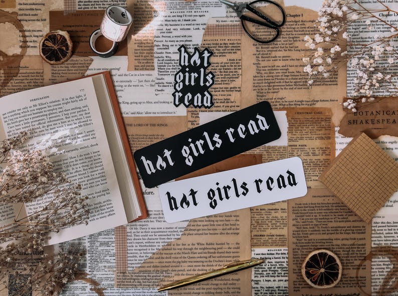 May include: Two black and white bookmarks with the text "hot girls read" in a gothic font. The bookmarks are on a table with a book, dried flowers, and other decorative items.