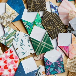 K&ouml;nnte beinhalten: Eine Sammlung kleiner, dekorativer Umschl&auml;ge mit verschiedenen Mustern und Farben, darunter florale, geometrische und abstrakte Designs. Die Umschl&auml;ge sind aus Papier und haben ein wei&szlig;es Innenleben.