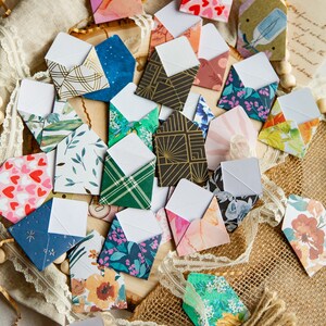 K&ouml;nnte beinhalten: Eine Sammlung kleiner, bunter Umschl&auml;ge mit verschiedenen Mustern und Designs. Die Umschl&auml;ge sind aus Papier und haben ein wei&szlig;es Innenleben. Sie sind auf einer Jutestoffoberfl&auml;che mit Spitze und Holzperlen angeordnet.