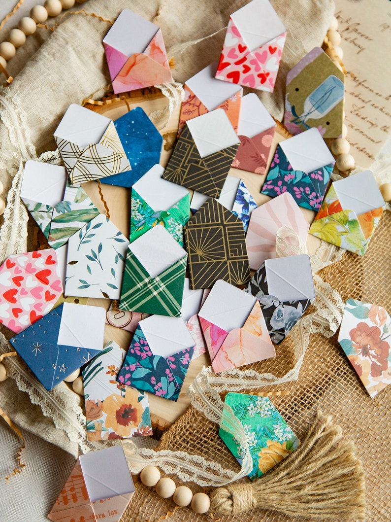 K&ouml;nnte beinhalten: Eine Sammlung kleiner, bunter Umschl&auml;ge mit verschiedenen Mustern und Designs. Die Umschl&auml;ge sind aus Papier und haben ein wei&szlig;es Innenleben. Sie sind auf einer Jutestoffoberfl&auml;che mit Spitze und Holzperlen angeordnet.