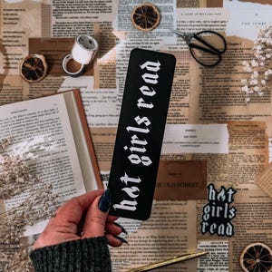 May include: A black bookmark with white text that reads "hot girls read". The bookmark is laying on a table with a book, a pair of scissors, and other decorative items.
