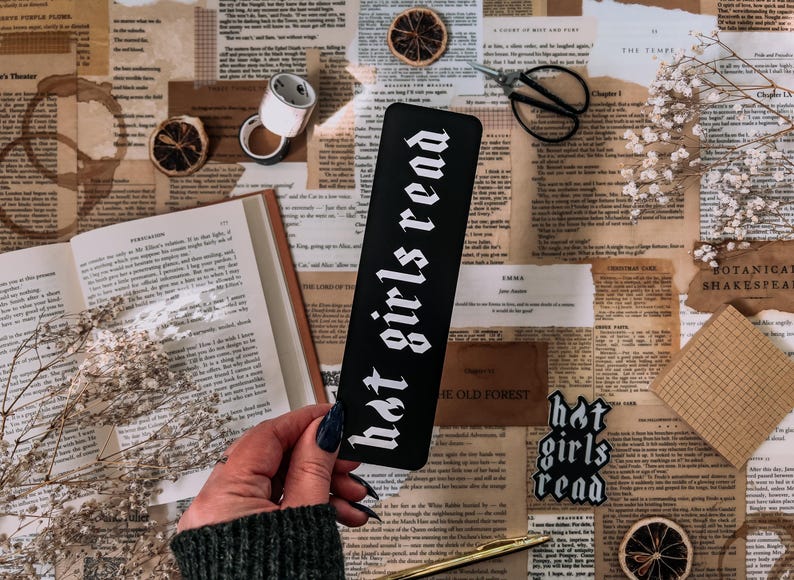 May include: A black bookmark with white text that reads "hot girls read". The bookmark is laying on a table with a book, a pair of scissors, and other decorative items.