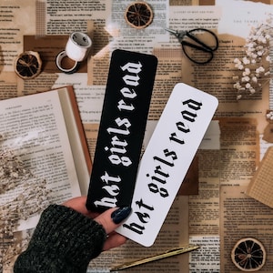 May include: Two black and white bookmarks with the text "hot girls read" in a gothic font. The bookmarks are on a table with a book, flowers, and other decorative items.