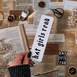 May include: A white bookmark with black gothic lettering that reads "hot girls read". The bookmark is on a table with a book, flowers, and other decorative items.