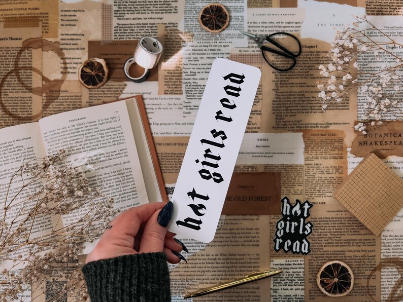 May include: A white bookmark with black gothic lettering that reads "hot girls read". The bookmark is on a table with a book, flowers, and other decorative items.