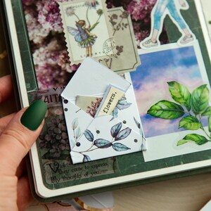 K&ouml;nnte beinhalten: Ein wei&szlig;er Umschlag mit floralem Muster und schwarzen Punkten. Der Umschlag ist ge&ouml;ffnet und ein kleines St&uuml;ck Papier mit dem Wort "flowers" darauf ist im Inneren sichtbar. Der Umschlag liegt auf einer gr&uuml;nen Oberfl&auml;che mit anderen dekorativen Elementen.