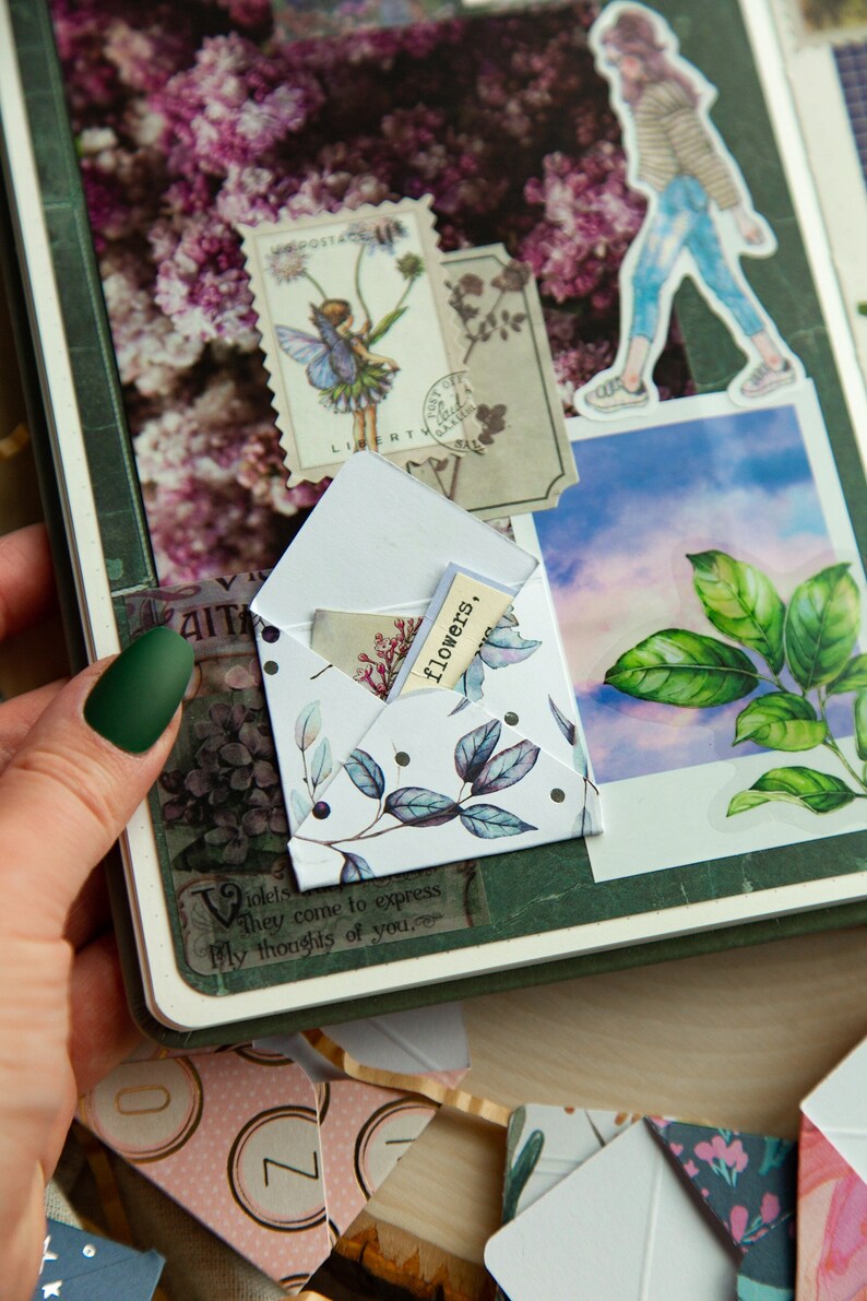 K&ouml;nnte beinhalten: Ein wei&szlig;er Umschlag mit floralem Muster und schwarzen Punkten. Der Umschlag ist ge&ouml;ffnet und ein kleines St&uuml;ck Papier mit dem Wort "flowers" darauf ist im Inneren sichtbar. Der Umschlag liegt auf einer gr&uuml;nen Oberfl&auml;che mit anderen dekorativen Elementen.