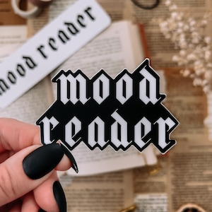 Könnte beinhalten: Ein schwarz-weißer Aufkleber mit den Worten "mood reader" in einer gotischen Schriftart. Der Aufkleber wird von einer Person mit schwarzem Nagellack gehalten. Weitere Elemente im Bild sind ein Lesezeichen, eine Schere und Trockenblumen.