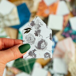 K&ouml;nnte beinhalten: Ein wei&szlig;er Umschlag mit einem floralen Muster in Graut&ouml;nen und Schwarz. Der Umschlag ist in der Mitte gefaltet und hat eine spitze Oberseite.