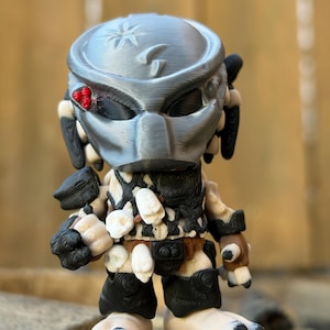 May include: A detailed figurine of a Predator character, featuring a silver helmet with a red eye, and a detailed body with black, white, and tan accents. The figure has a unique design with intricate details, showcasing a blend of colors and textures.