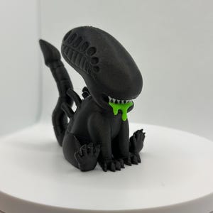 Puede incluir: Una figura de extraterrestre negra impresa en 3D con una sustancia verde que gotea de su boca. El extraterrestre est&aacute; sentado en cuclillas con la cola enroscada detr&aacute;s de &eacute;l.
