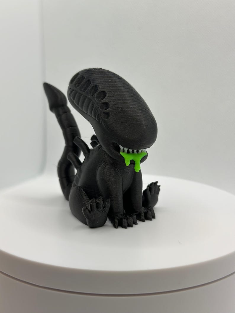 Puede incluir: Una figura de extraterrestre negra impresa en 3D con una sustancia verde que gotea de su boca. El extraterrestre est&aacute; sentado en cuclillas con la cola enroscada detr&aacute;s de &eacute;l.