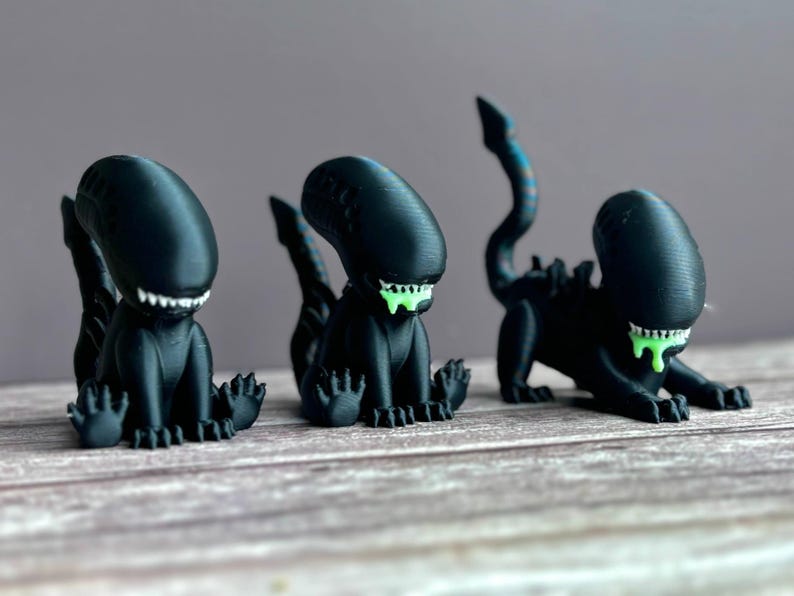 Puede incluir: Tres figuras de alien&iacute;genas negras con verde goteando de sus bocas. Los alien&iacute;genas est&aacute;n en posici&oacute;n sentada con sus colas enroscadas detr&aacute;s de ellos.
