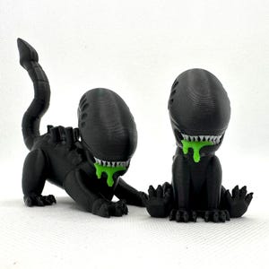 Puede incluir: Dos figuras negras impresas en 3D de una criatura extraterrestre con una cola larga y baba verde goteando de su boca.