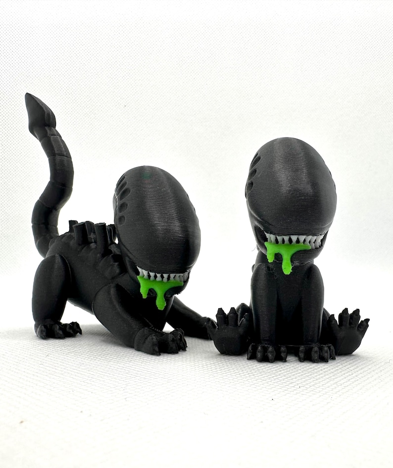 Puede incluir: Dos figuras negras impresas en 3D de una criatura extraterrestre con una cola larga y baba verde goteando de su boca.
