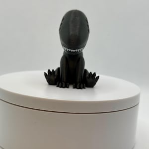 Puede incluir: Una peque&ntilde;a figura de dinosaurio negra impresa en 3D sentada sobre una base circular blanca. El dinosaurio tiene una sonrisa llena de dientes y est&aacute; sentado con las piernas cruzadas.