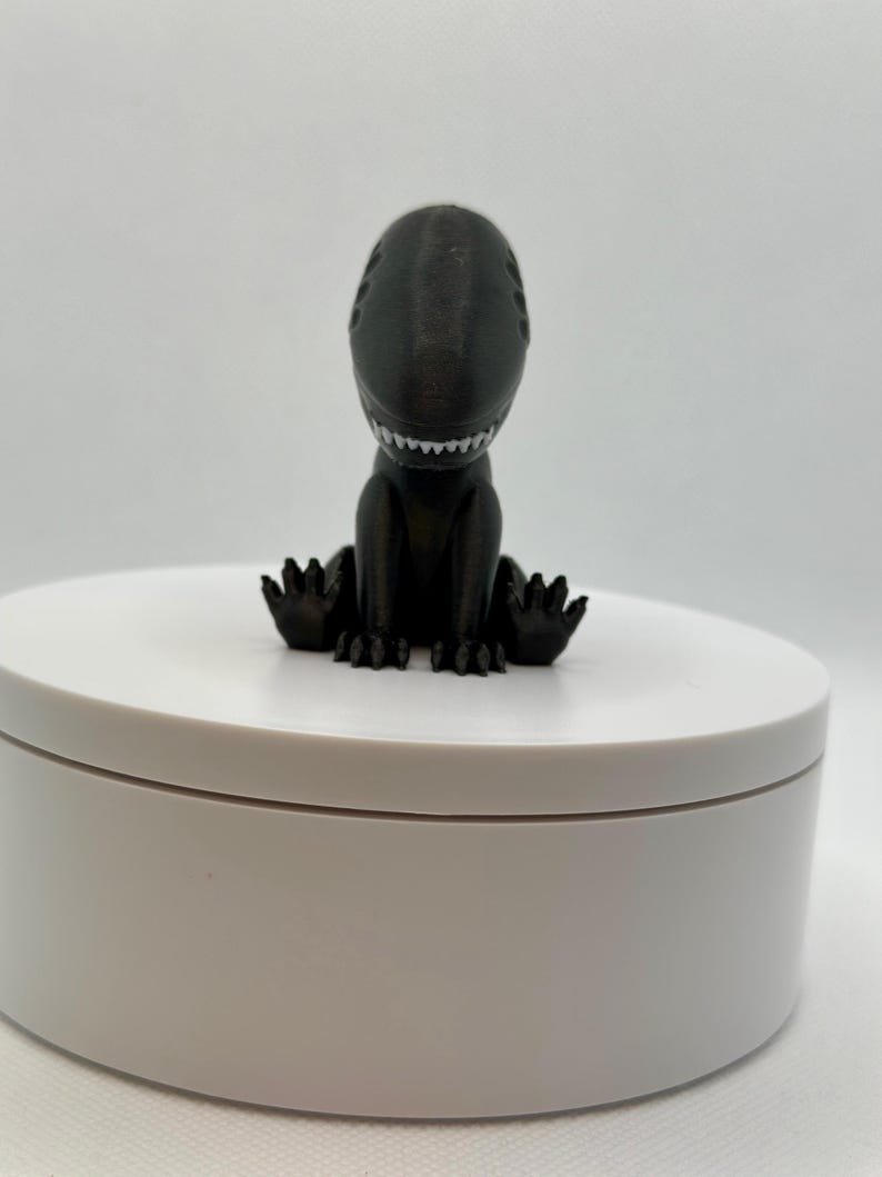 Puede incluir: Una peque&ntilde;a figura de dinosaurio negra impresa en 3D sentada sobre una base circular blanca. El dinosaurio tiene una sonrisa llena de dientes y est&aacute; sentado con las piernas cruzadas.
