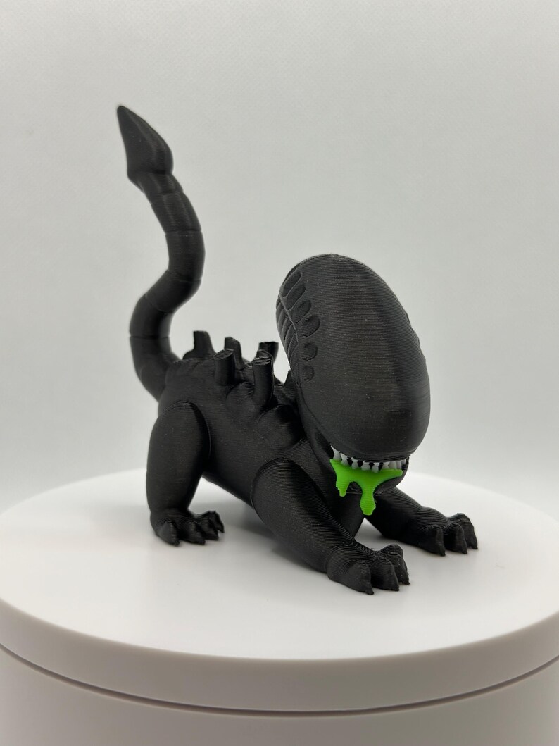 Puede incluir: Una figura de criatura extraterrestre negra impresa en 3D con una cola larga y baba verde goteando de su boca.