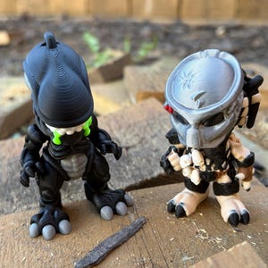 May include: Two 3D-printed figurines: a black Xenomorph with green accents and a Predator with silver and tan details. The figures are posed on a wooden surface, showcasing their detailed designs and iconic features.