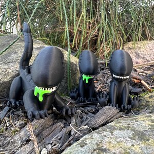 Puede incluir: Tres figuras negras impresas en 3D de una criatura extraterrestre con baba verde goteando de su boca. Las figuras est&aacute;n en posici&oacute;n sentada con las piernas cruzadas. Las figuras est&aacute;n sobre una cama de astillas de madera y rocas.