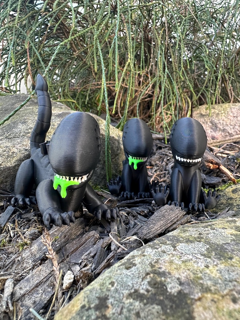 Puede incluir: Tres figuras negras impresas en 3D de una criatura extraterrestre con baba verde goteando de su boca. Las figuras est&aacute;n en posici&oacute;n sentada con las piernas cruzadas. Las figuras est&aacute;n sobre una cama de astillas de madera y rocas.